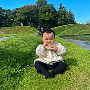 Grace is registered to the contest to win money with this photo: baby, child, grass, lawn, pond, water, sky, blue_sky, trees, greenery, sunny, outdoor, park, sitting, headband, clothing, stroller, people, nature, chewing