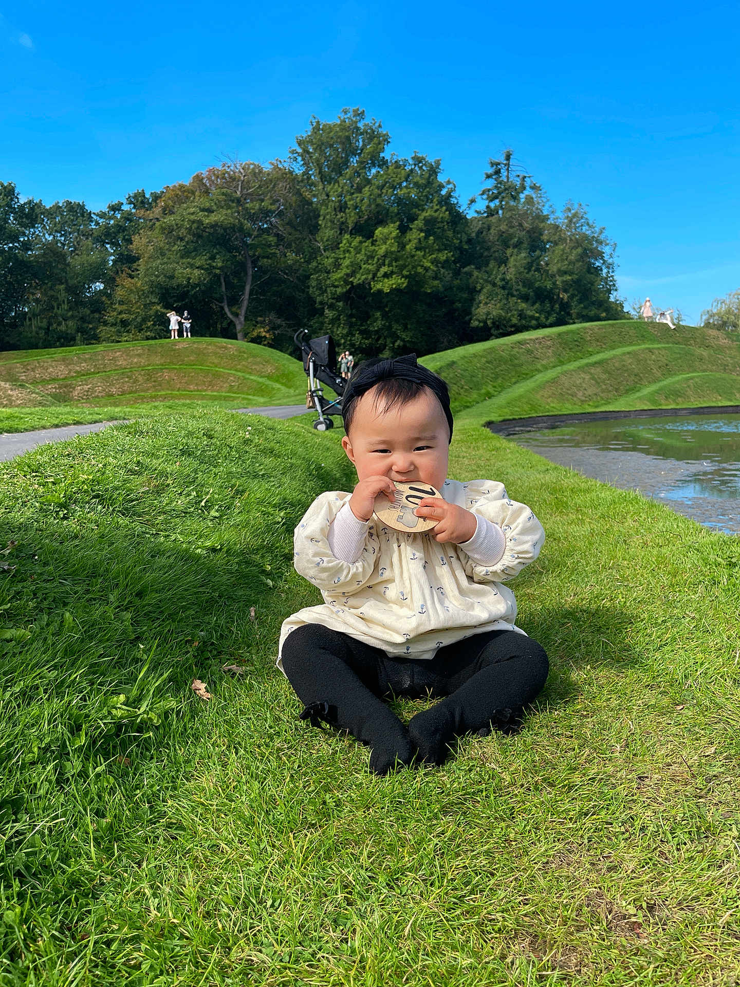 Grace is registered to the contest to win money with this photo: baby, child, grass, lawn, pond, water, sky, blue_sky, trees, greenery, sunny, outdoor, park, sitting, headband, clothing, stroller, people, nature, chewing
