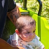 baby, child, infant, outdoor, sunlight, greenery, playground, seated, adult, holding, face, portrait, curious, short_hair, bib, clothing, cartoon_print, green_seat, tattoo, family