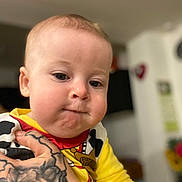 Maël a rejoint le concours — aidez-le/la à gagner de superbes lots ! baby, child, tattoo, hand, yellow_clothing, cow_print, portrait, indoor, closeup, cute, infant, person, skin, expression, holding, face, young, childhood, blurred_background, colorful_clothing