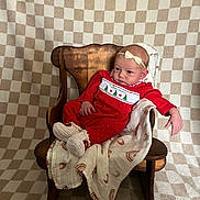 Kristina is registered to the contest to win money with this photo: baby, infant, red_clothing, christmas_theme, wooden_chair, blanket, headband, patterned_background, indoors, portrait, cute, holiday, cozy, smiling, sitting, newborn, warm, decor, soft_lighting, relaxed