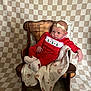 baby, infant, red_clothing, christmas_theme, wooden_chair, blanket, headband, patterned_background, indoors, portrait, cute, holiday, cozy, smiling, sitting, newborn, warm, decor, soft_lighting, relaxed