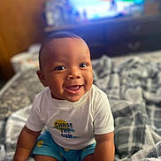Roman joined the competition — help win amazing prizes! baby, bed, blanket, blue_shorts, child, clothing, cozy, cute, face, happy, home, indoor, infant, person, portrait, sitting, smiling, soft_light, warm, white_shirt