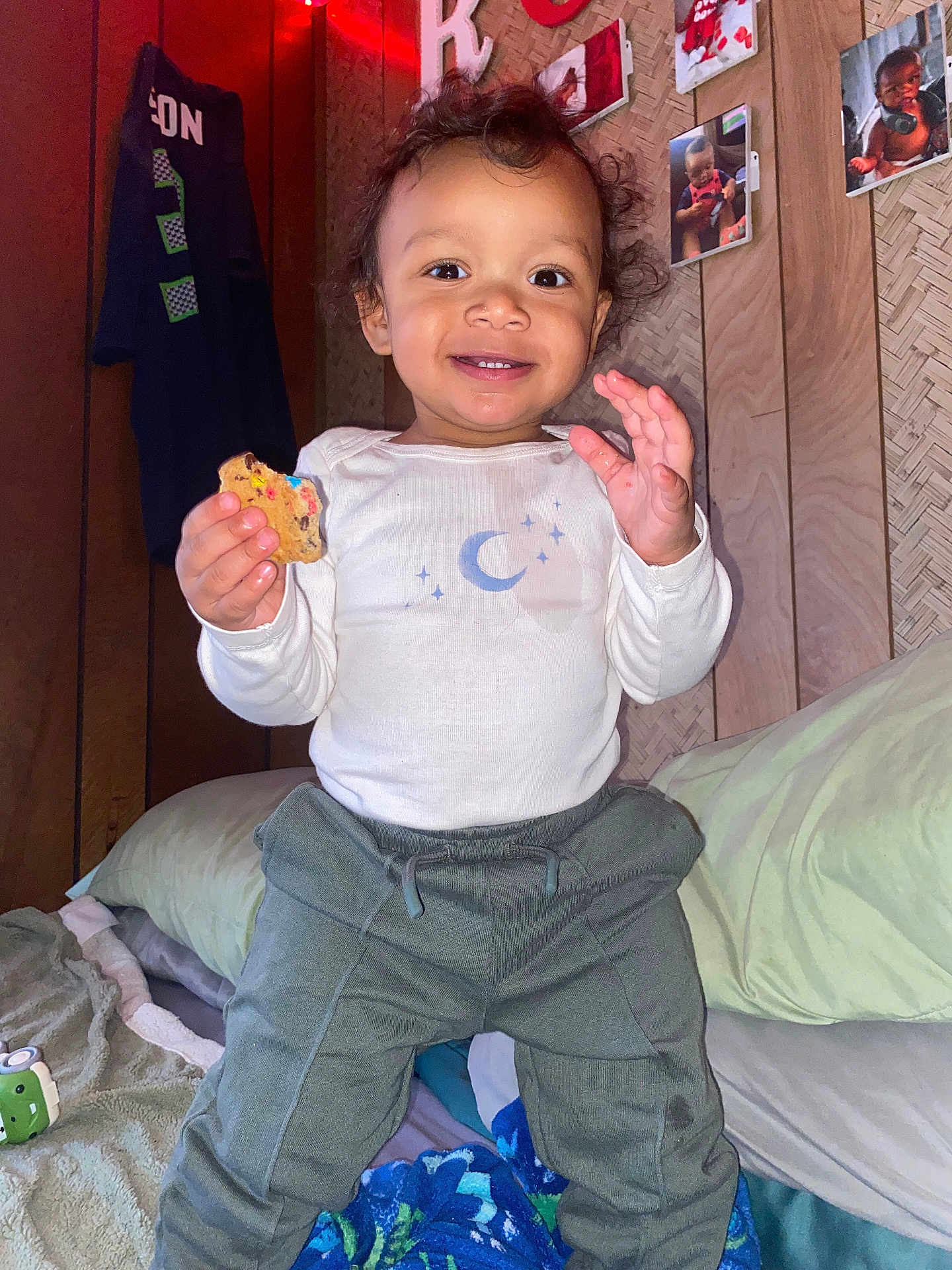 Roman is registered to the contest to win money with this photo: child, toddler, baby, smiling, cookie, onesie, pants, pillow, blanket, toy, wall_photos, wood_paneling, face, hands, indoor, bed, cozy, portrait, flash, happy
