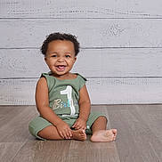 Cyrus joined the competition — help win amazing prizes! baby, child, birthday, smiling, green_outfit, sitting, floor, wooden_floor, white_background, portrait, cute, happy, one_year_old, indoors, infant, person, celebration, clothing, toes, curly_hair