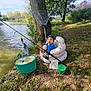 toddler, inflatable_chair, fishing_rod, fish, lake, tree, grass, bucket, child, outdoor, nature, water, fishing_net, cap, blue_jacket, sunlight, leaves, fishing, container, recreational