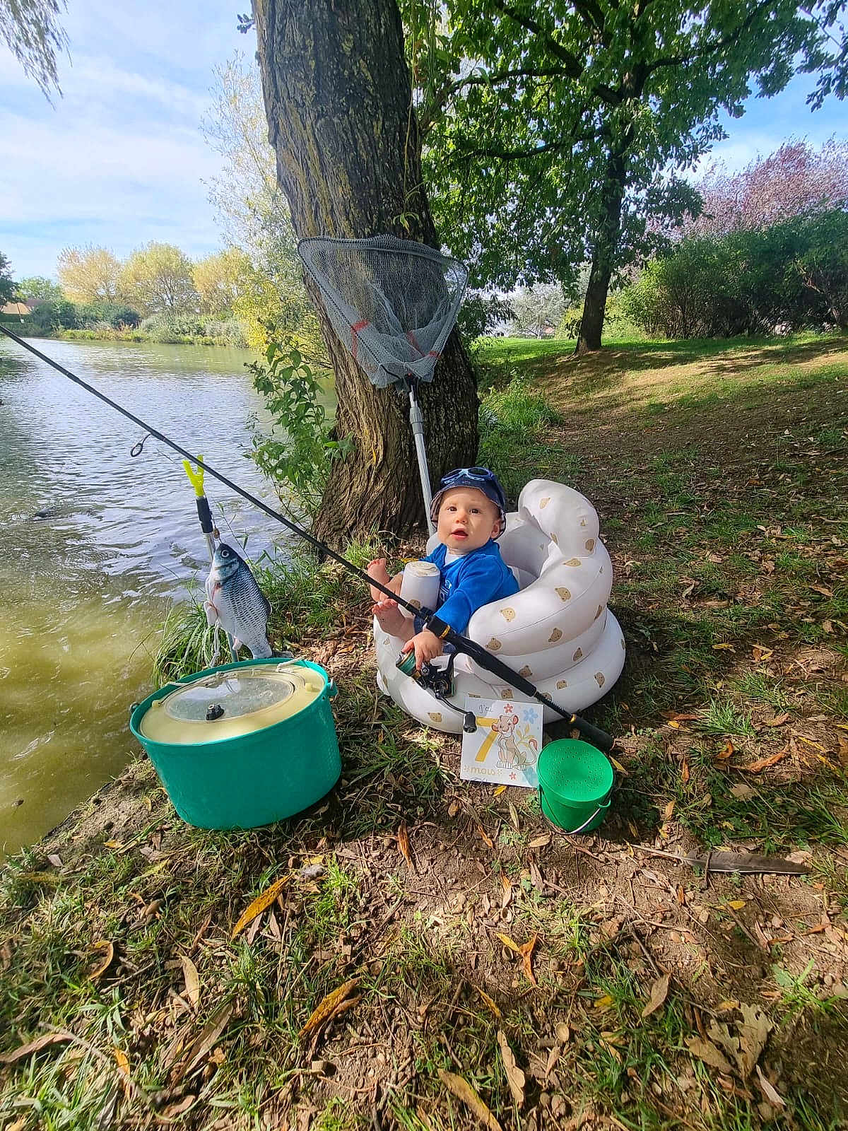 Timéo participe au concours pour gagner de l'argent avec cette photo : toddler, inflatable_chair, fishing_rod, fish, lake, tree, grass, bucket, child, outdoor, nature, water, fishing_net, cap, blue_jacket, sunlight, leaves, fishing, container, recreational