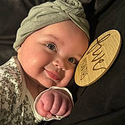Amalia is registered to the contest to win money with this photo: baby, infant, headwrap, smile, face, fist, wooden_sign, clothing, closeup, portrait, cute, child, soft_lighting, cozy, warm, skin, expression, sleepwear, hand, background