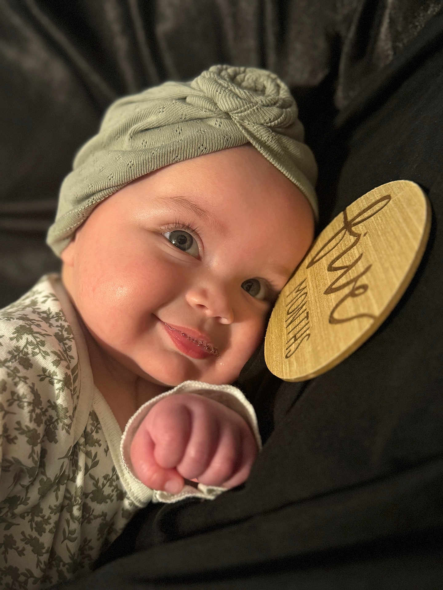 Amalia is registered to the contest to win money with this photo: baby, infant, headwrap, smile, face, fist, wooden_sign, clothing, closeup, portrait, cute, child, soft_lighting, cozy, warm, skin, expression, sleepwear, hand, background
