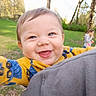 baby, child, smiling, outdoor, grass, trees, person, blue_eyes, yellow_clothing, happy, face, nature, greenery, playful, blurred_background, casual_clothing, holding_camera, joyful, park, family
