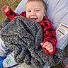 baby, child, smiling, red_jacket, gray_blanket, baby_seat, bottle, outdoor, happy, face, hand, clothing, footwear, plaid, infant, person, cozy, blanket, seat, cute