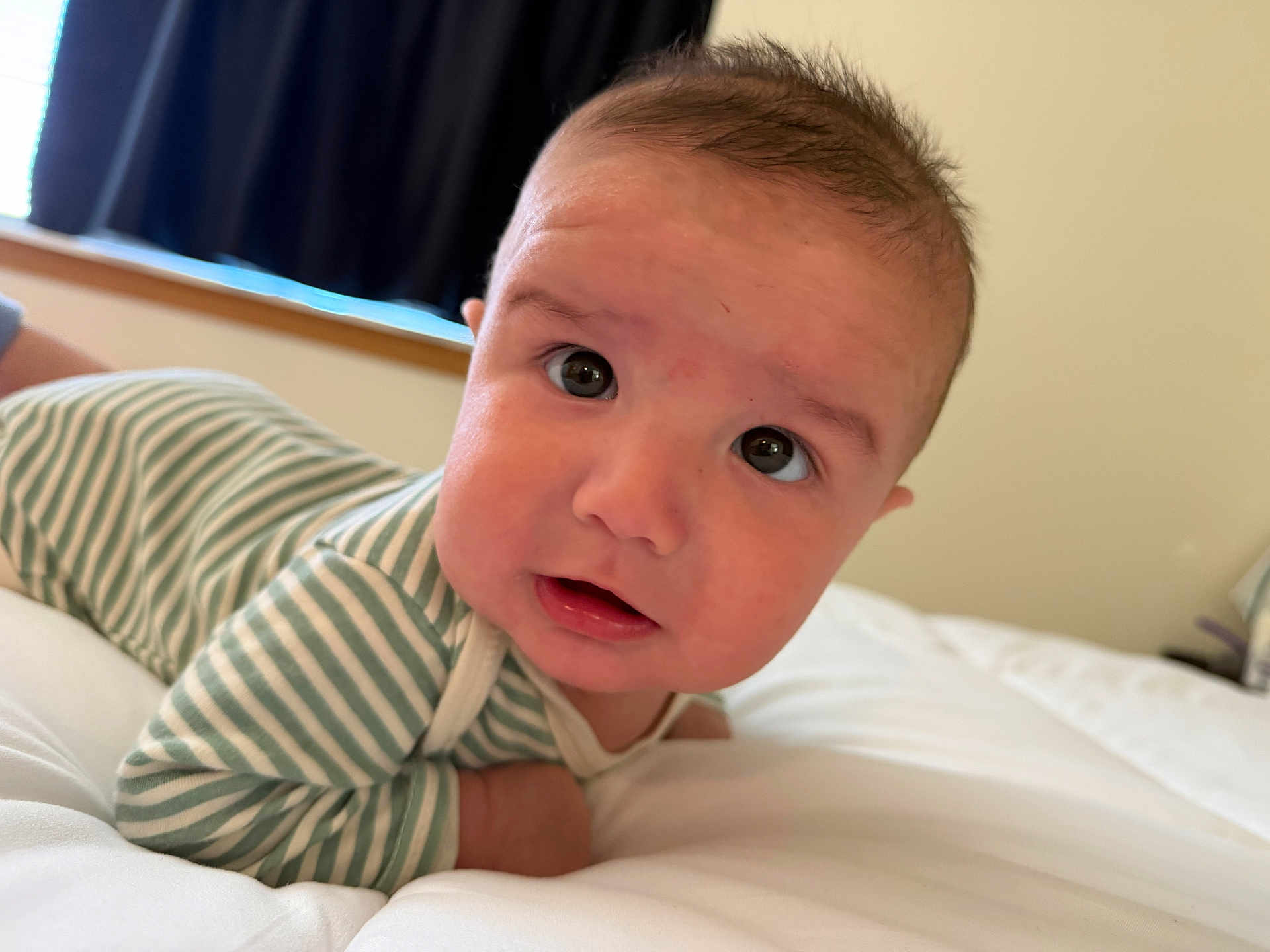 Damian is registered to the contest to win money with this photo: baby, infant, face, eyes, mouth, striped_clothing, bed, white_bed, soft, curious, indoors, portrait, closeup, head, skin, person, young_child, sleepwear, blanket, window