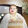 Abby is registered to the contest to win money with this photo: baby, basket, candle, child, curious_expression, cute, decor, gold_accessory, greenery, hands, headpiece, indoor, infant, plant, portrait, sitting, smile, white_clothing, wooden_background, woven_texture