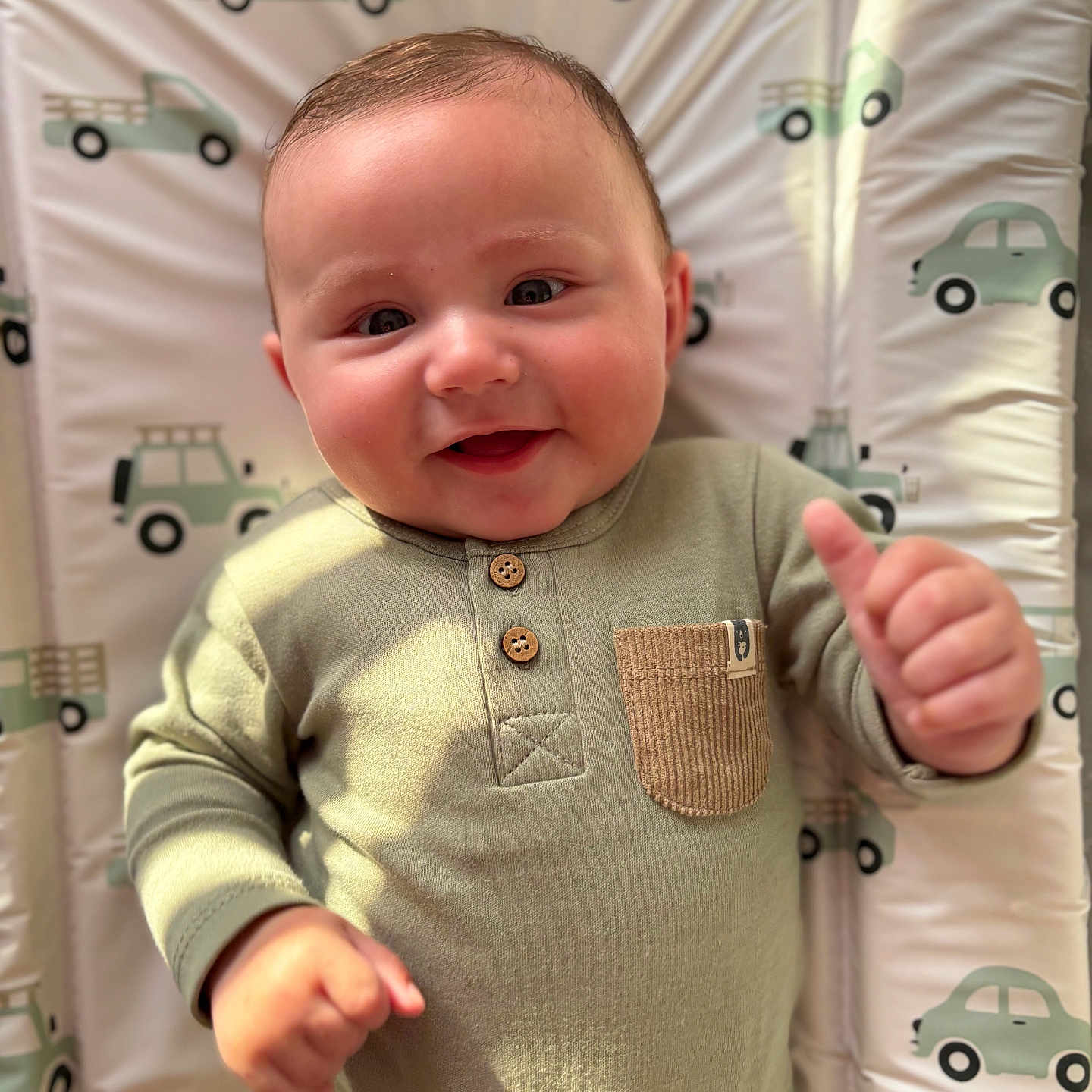 Harrison is registered to the contest to win money with this photo: baby, baby_gesture, car, child, cute, face, green_clothing, hand, happy, indoors, infant, patterned_mat, pocket, portrait, smiling, soft_lighting, thumbs_up, truck, vehicle_pattern, wooden_buttons
