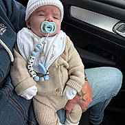 Jesse is registered to the contest to win money with this photo: baby, infant, pacifier, beanie, knitwear, bib, car_interior, adult_hand, tattoo, jeans, socks, seated, close_up, person, clothing, comfort, transportation, cute, indoor, portrait