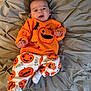 baby, infant, smiling, orange_clothing, pumpkin_pattern, halloween, blanket, bed, cute, happy, child, person, pajamas, festive, cozy, indoor, soft_fabric, tufted, white_socks, casual