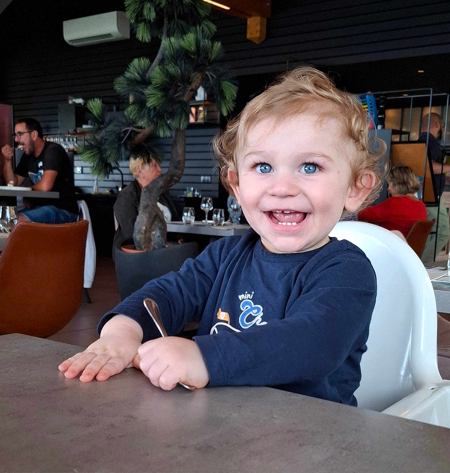 Maylon a rejoint le concours — aidez-le/la à gagner de superbes lots ! toddler, child, high_chair, smiling, blue_eyes, curly_hair, indoor, restaurant, table, spoon, happy, person, casual_clothing, seated, background_people, plant, modern_interior, furniture, light, portrait