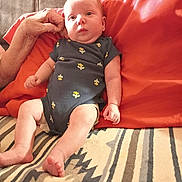 Serenity is registered to the contest to win money with this photo: baby, infant, child, portrait, sitting, lying_down, wide_eyed, indoors, red_pillows, blanket, cactus_print, onesie, adult_arm, furniture, couch, textile, cozy, home, patterned_blanket, photography