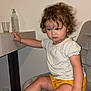 barefoot, bottle, bracelet, chair, child, curly_hair, expression, floor, furniture, glass, indoor, person, room, sitting, skin, table, toddler, wall, white_shirt, yellow_shorts
