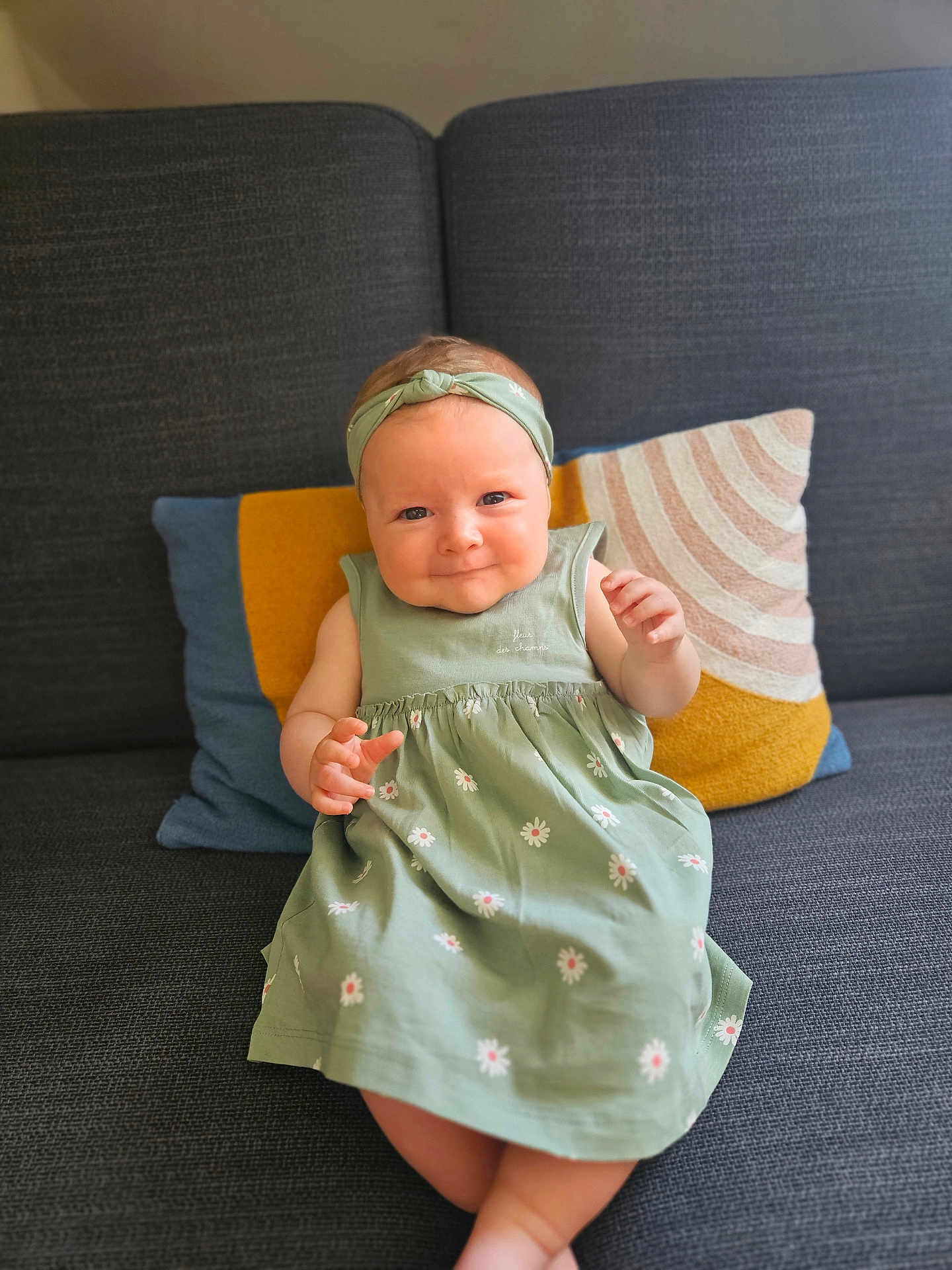 Cassie participe au concours pour gagner de l'argent avec cette photo : baby, child, dress, headband, couch, pillow, smile, sitting, indoors, cute, green, floral_pattern, happy, portrait, infant, clothing, face, person, home, comfort