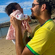 Anaïa a rejoint le concours — aidez-le/la à gagner de superbes lots ! baby, man, beach, sky, clouds, sand, water, holding, smiling, yellow_shirt, pink_dress, happy, portrait, outdoor, family, love, glasses, short_hair, casual, sunny