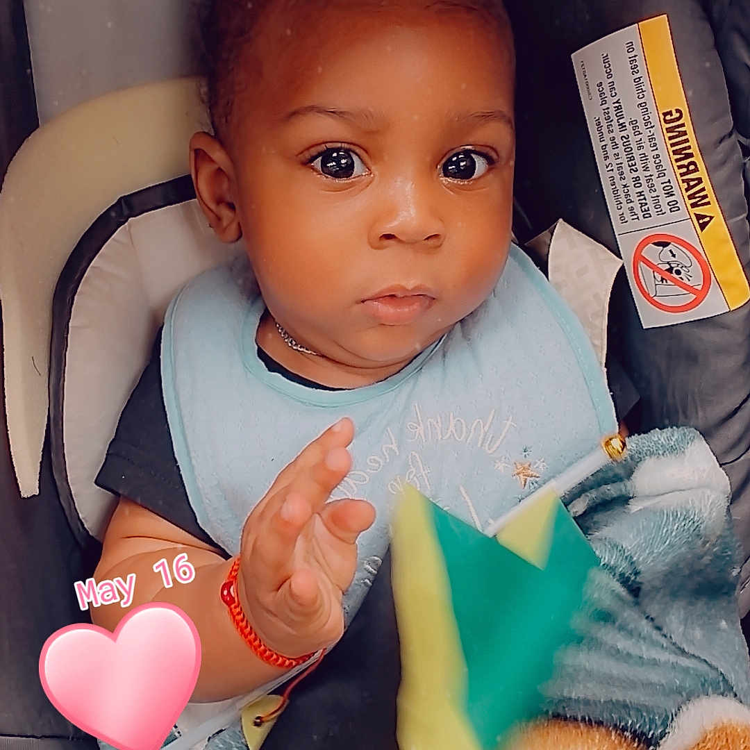 Jadane is registered to the contest to win money with this photo: baby, child, stroller, bib, blanket, toy, star, hand, face, expression, curious, seated, red_bracelet, clothing, indoor, infant, portrait, cute, person, pink_heart