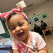 Evangeline is registered to the contest to win money with this photo: baby, child, laughing, pink_headband, hello_kitty, clapping, indoor, waiting_room, couch, adults, woman, phone, wood_floor, ceiling_lights, wall_art, happy, person, casual_clothing, smile, portrait