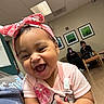 baby, child, laughing, pink_headband, hello_kitty, clapping, indoor, waiting_room, couch, adults, woman, phone, wood_floor, ceiling_lights, wall_art, happy, person, casual_clothing, smile, portrait