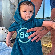 Hector participe au concours pour gagner de l'argent avec cette photo : baby, child, towel, hooded_towel, beach, sand, tattoo, adult_hand, building, window, stone_wall, blue_clothing, outdoor, person, standing, skin, daylight, expression, holding, foot
