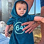 baby, child, towel, hooded_towel, beach, sand, tattoo, adult_hand, building, window, stone_wall, blue_clothing, outdoor, person, standing, skin, daylight, expression, holding, foot
