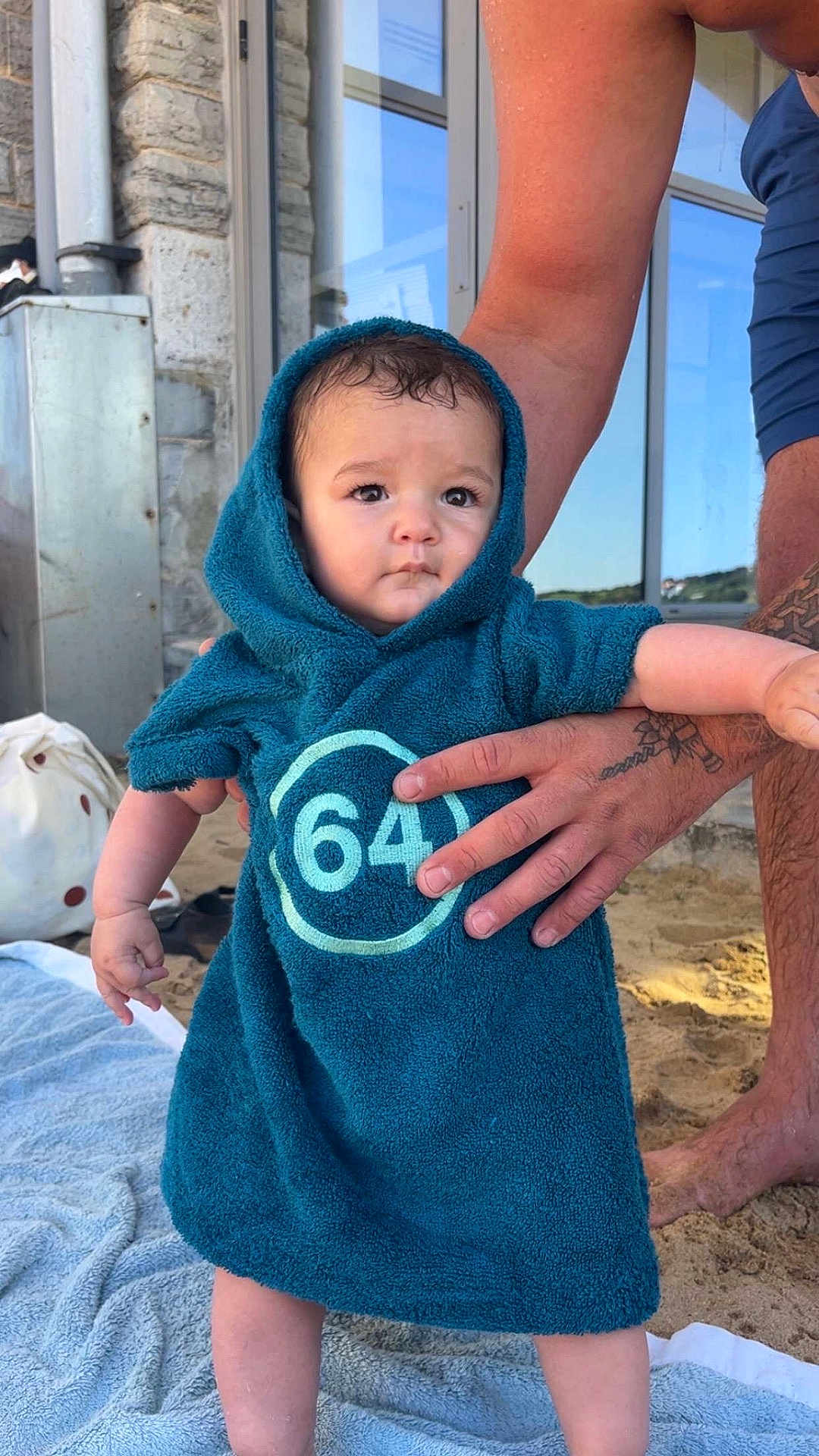 Hector participe au concours pour gagner de l'argent avec cette photo : baby, child, towel, hooded_towel, beach, sand, tattoo, adult_hand, building, window, stone_wall, blue_clothing, outdoor, person, standing, skin, daylight, expression, holding, foot