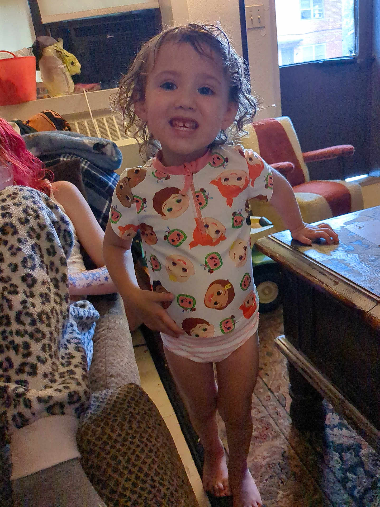 Eliza is registered to the contest to win money with this photo: child, indoor, smile, curly_hair, colorful_clothing, table, person, furniture, window, carpet, blanket, seated_person, leopard_print, living_room, toy, casual_pose, barefoot, happy, young_child