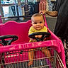 baby, shopping_cart, pink, child, steering_wheel, indoor, store, tattoo, hand, person, floor, clothing, window, door, product_stack, car_cart, foot, metal, plastic, curious