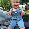 Aurora is registered to the contest to win money with this photo: baby, child, denim_overalls, smiling, happy, outdoor, car, black_car, hood, green_trees, suburban, nature, sunlight, cute, portrait, person, casual_clothing, shorts, grass, daytime