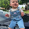 baby, child, denim_overalls, smiling, outdoor, car, hood, trees, greenery, sunlight, cute, happy, person, clothing, face, shorts, nature, daylight, infant, blue