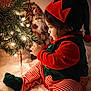 toddler, child, christmas_tree, christmas_lights, christmas_ornaments, elf_costume, holiday, festive, decorations, indoor, seasonal, toy, red_and_green, striped_pants, hat, cute, portrait, warm_lighting, celebration, santa_figurine