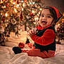 baby, child, smiling, holiday, christmas_tree, christmas_ornaments, elf_costume, red_and_green, striped_pants, festive, indoors, happy, clapping, blanket, decorations, cute, portrait, celebration, seasonal, warm_lighting