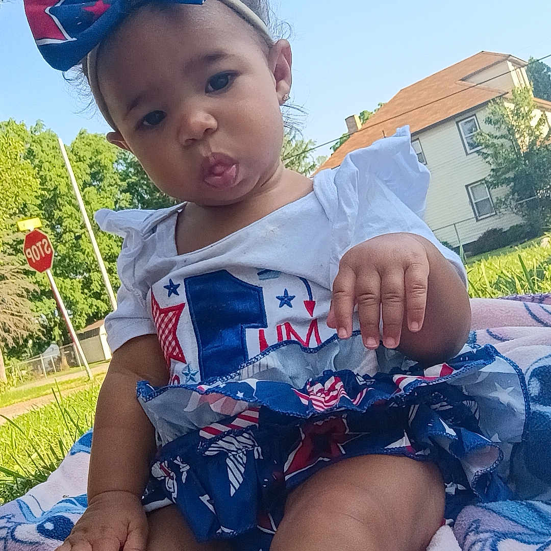 Lilianne joined the competition — help win amazing prizes! blanket, bow, child, cute, daytime, feet, grass, headband, house, legs, outdoor, person, playful, shirt, skirt, stop_sign, sunny, toddler, tongue, trees