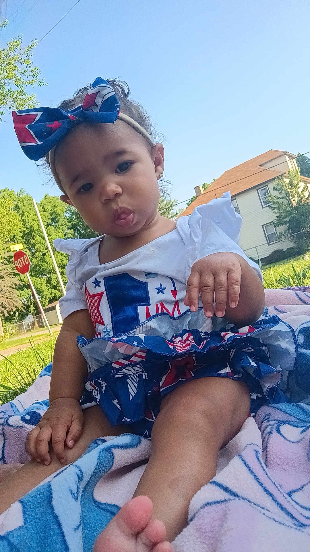 Lilianne joined the competition — help win amazing prizes! toddler, child, outdoor, grass, blanket, headband, bow, tongue, playful, shirt, skirt, stop_sign, house, trees, sunny, daytime, person, legs, feet, cute