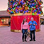 couple, dog, christmas_tree, holiday_decorations, lights, ornaments, outdoor, evening, pavement, retail_stores, woman, man, leash, casual_clothing, boots, glasses, palm_trees, shopping_center, festive, smiling