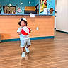 child, toddler, smiling, portrait, indoor, reception_desk, clinic, waiting_room, beach_ball, toy, baseball_cap, striped_shirt, shorts, sneakers, wood_floor, colorful, flowers, fruit_artwork, stuffed_animal, happy