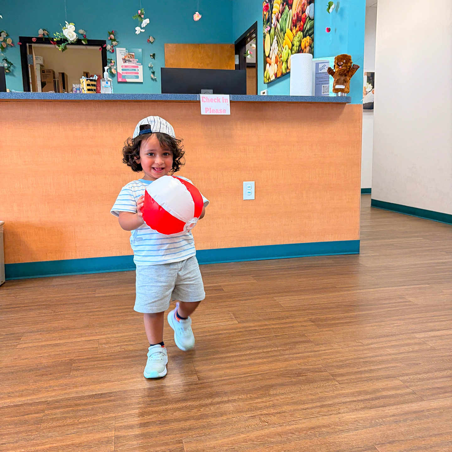Alaya joined the competition — help win amazing prizes! child, toddler, smiling, portrait, indoor, reception_desk, clinic, waiting_room, beach_ball, toy, baseball_cap, striped_shirt, shorts, sneakers, wood_floor, colorful, flowers, fruit_artwork, stuffed_animal, happy