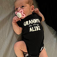 Arya is registered to the contest to win money with this photo: newborn, baby, infant, child, portrait, pacifier, onesie, black_clothing, grandma, text_on_clothing, lying_down, bed, blanket, socks, striped_socks, indoor, face, hands, feet, adorable