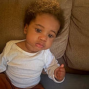 Elijah joined the competition — help win amazing prizes! toddler, child, curly_hair, white_shirt, brown_pants, couch, sofa, indoor, seated, person, face, expression, young_child, casual_clothing, home, furniture, portrait, skin, hand, relaxed