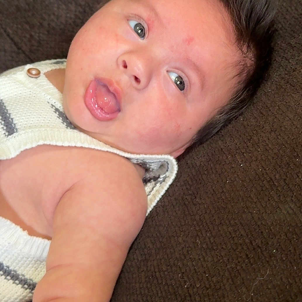 Jose is registered to the contest to win money with this photo: baby, infant, face, tongue_out, arm, knitted_clothing, couch, expression, red_bracelet, skin, eyes, hair, lying_down, comfortable, cute, indoors, texture, closeup, person, child