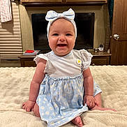 Tatum is registered to the contest to win money with this photo: baby, infant, child, smiling, headband, bow, dress, daisy_pattern, sitting, blanket, bed, bedroom, television, tv_screen, cabinet, wooden_furniture, portrait, happy, indoor, cute