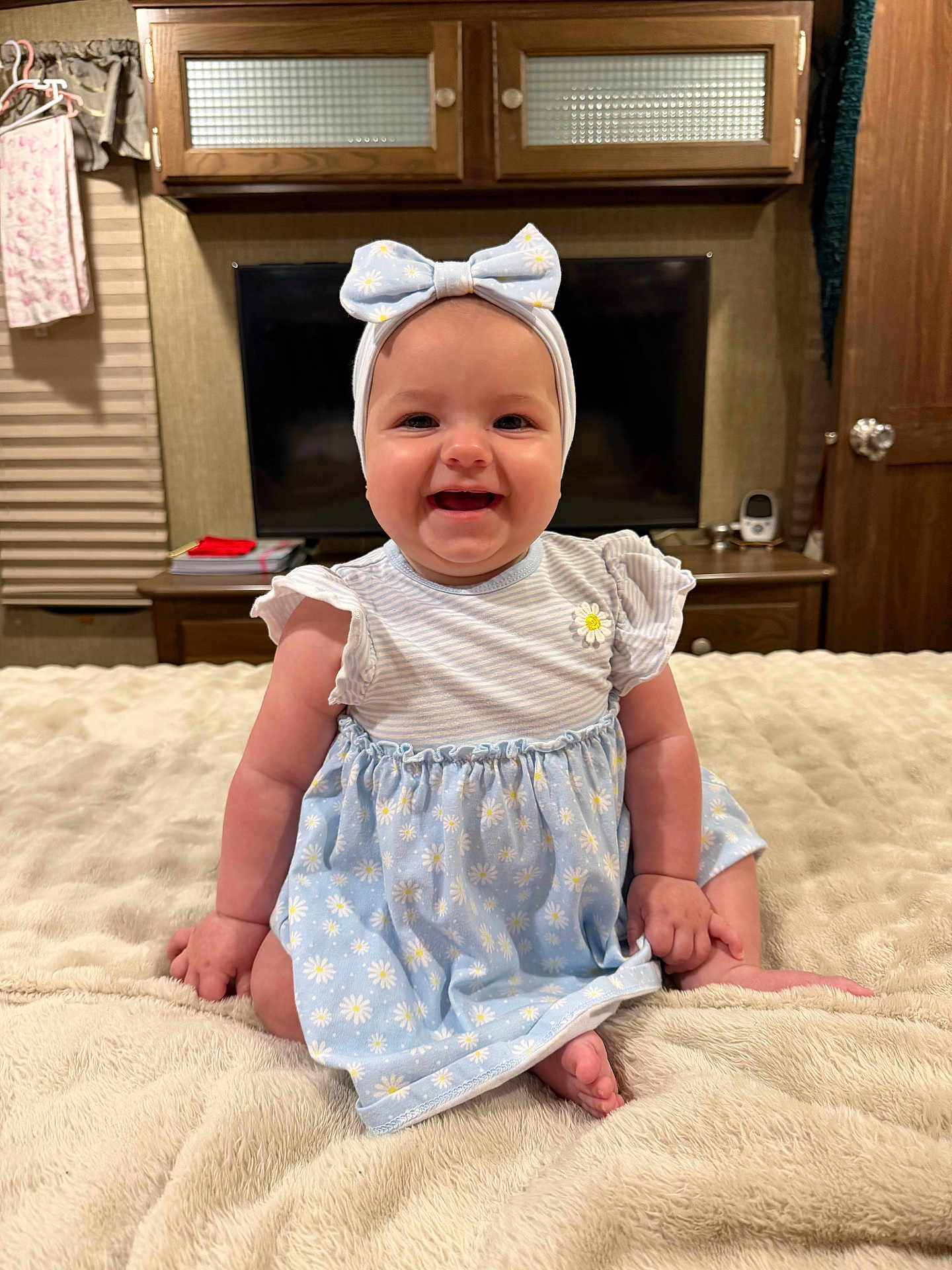 Tatum is registered to the contest to win money with this photo: baby, infant, child, smiling, headband, bow, dress, daisy_pattern, sitting, blanket, bed, bedroom, television, tv_screen, cabinet, wooden_furniture, portrait, happy, indoor, cute