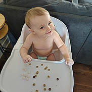 Isabella is registered to the contest to win money with this photo: baby, high_chair, cereal, smile, indoor, child, diaper, furniture, living_room, table, wood_floor, person, face, young_child, seat, playful, cute, happy, messy, home