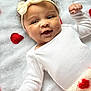 Daylee is registered to the contest to win money with this photo: baby, infant, smile, headband, bow, white_clothing, blanket, red_rose_petals, soft_texture, lying_down, cute, happy, portrait, closeup, child, warm, cozy, indoor, newborn, adorable