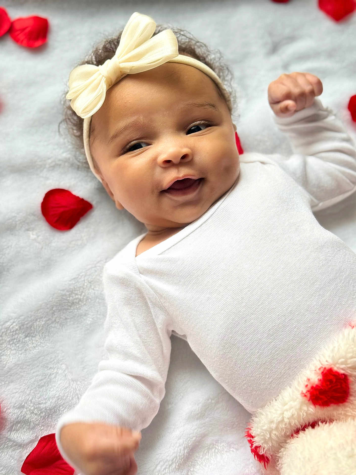 Daylee is registered to the contest to win money with this photo: baby, infant, smile, headband, bow, white_clothing, blanket, red_rose_petals, soft_texture, lying_down, cute, happy, portrait, closeup, child, warm, cozy, indoor, newborn, adorable